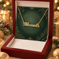 A gold script affirmation necklace reading 'Blessed' with a small round charm, displayed on a dark green decorative card inside an open red gift box. The scene is styled with holiday lights, pine branches, and gold ornaments in the background.