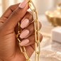 A close-up of a hand with nude pink manicured nails holding a chunky gold chain necklace with alternating large and small links, some embellished with clear crystals.