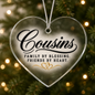 A heart-shaped clear glass Christmas ornament hanging from a silver string, printed with the words 'Cousins Family by Blessing. Friends by Heart.