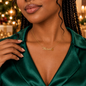 A close-up of a woman wearing a delicate gold necklace with the word 'Blessed' in cursive script and a small ornament at the end, styled with a dark green satin top against a festive holiday background.