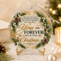 Round clear glass Christmas ornament on a small gold stand, printed with a festive wreath design, pinecones, white berries, and the message 'You Are Forever A Part of Every Christmas.' The ornament is photographed on a marble surface with holiday decor and warm blurred lights in the background.