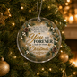 A round clear Christmas ornament hanging on a lit Christmas tree, printed with a wreath design, pinecones, berries, and the message, 'You are FOREVER a PART of every Christmas.'.
