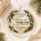 A round clear Christmas ornament with printed holiday greenery, gold foil details, and the message 'You Are Forever A Part of Every Christmas,' shown on a cream fabric surface with pine branches and a wrapped gift nearby.