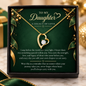 A gold heart-shaped pendant necklace with a clear central gemstone, presented inside a black gift box with a dark green card addressed to a daughter. The card features elegant gold text and holiday-themed gold and green leaf illustrations.