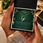 A person holds an open black gift box containing a heart-shaped silver pendant necklace on a dark green sentimental card addressed 'To My Daughter.' The scene is styled with hands, holiday greenery, and warm blurred background lights, so this is Lifestyle rather than Product/Packaging.