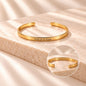A gold open cuff bracelet with engraved text, hold on to hope on the outside displayed on a beige stone slab with soft shadows. A circular inset shows the reverse side with the engraved phrase 'Hold on to yourself'.