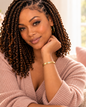 A woman with long, tight, dark brown box braids sits indoors, wearing a dusty pink ribbed cardigan and a gold bracelet. The background shows a softly blurred living room setting with a window and pillows.