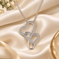A silver-toned double-heart pendant necklace paved with small clear stones, resting on draped champagne satin fabric with small white flowers and a gold ring partially visible in the background.