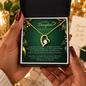 A person holds an open black gift box containing a gold heart-shaped pendant necklace with a clear rhinestone, displayed on a dark green sentimental card addressed 'To My Daughter'. The background is softly blurred with warm bokeh lights and a card reading 'Always Proud of You'.