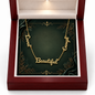 A gold nameplate necklace spelling 'Loved, 'Enough', and 'Beautiful' is displayed inside an open dark red presentation box with a green insert. The necklace is centered on the box, and the box interior is lit from above.
