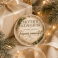 Round clear acrylic Christmas ornament with gold foil and green botanical accents, hanging in front of a wrapped gift box. The ornament reads “MOTHER & DAUGHTER,” “Two hearts.