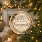 Round glass Christmas ornament hanging from a Christmas tree branch, featuring the printed message 'MOTHER & DAUGHTER' and 'Forever connected BY LOVE' with green foliage illustrations, set against a warm, blurred holiday living room background.