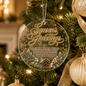 A round clear acrylic Christmas ornament hanging on a decorated tree, featuring gold foil-style script reading “Season’s Greetings,” a pinecone and evergreen branch design, and a warm printed holiday message. The ornament is shown in a real holiday tree setting with lights and other ornaments in the background.