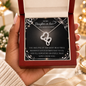 A hand holds an open red jewelry gift box containing a silver heart-link necklace displayed on a black sentimental card addressed to a daughter-in-law. The background is softly blurred with warm holiday lights and gift boxes.