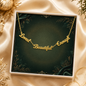 A gold script affirmation necklace reading 'Loved Beautiful Enough' displayed inside an open white gift box with a dark green velvet insert. The box is styled on a beige satin fabric with gold decorative accents.