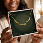 A gold script affirmation nameplate necklace reading 'Loved Beautiful Enough' displayed on a dark green insert inside a white gift box, held by a smiling woman in a softly lit indoor setting.