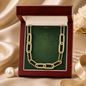 A gold paperclip chain necklace with pavé-set clear stones, displayed inside an open dark red wooden jewelry box with a green damask insert. The box is styled on a beige satin fabric background with a pearl and a white rose in a vase.